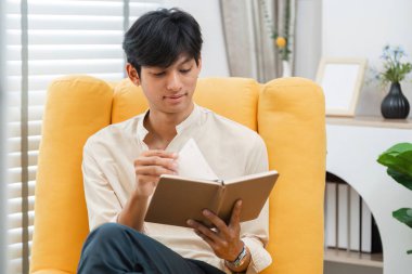 Genç bir adam sarı bir sandalyede rahat bir şekilde oturuyor, parlak, modern bir oturma odasında doğal ışık ve şık dekorasyonlu bir kitaptan keyif alıyor..