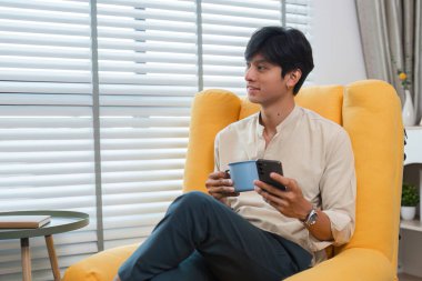 Genç bir adam modern bir evde rahatça oturur, bir fincan ve akıllı telefonla rahatlar, keyfine bakar..