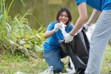 Mavi gömlekli gönüllüler parktan çöp topluyorlar, takım çalışması ve çevrenin korunmasına bağlılıklarını gösteriyorlar..