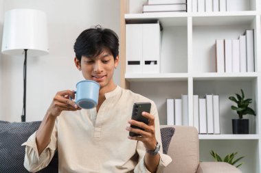 Genç bir adam, modern bir oturma odasında akıllı telefonunu karıştırırken bir fincan kahvenin tadını çıkarırken koltukta rahatça oturur..