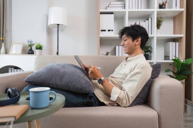 Genç bir adam rahat bir şekilde koltukta oturur, modern bir oturma odasında rahatlamak için tablet kullanır..