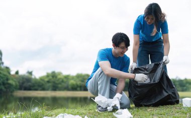 Mavi gömlekli ve eldivenli gönüllü bir çift parkta çöp topluyor, çevre koruma ve kamu hizmetini destekliyorlar..