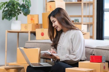 Evde çalışan genç bir kadın, online satış işini yönetiyor. Etrafı paketlerle çevrili ve dizüstü bilgisayar ve tablet kullanıyor..