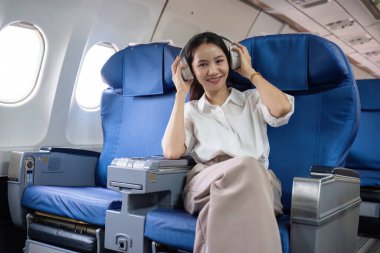 Kulaklık takan genç bir kadın, uçakta rahat bir şekilde otururken müzikten zevk alıyor. Seyahat ve yaşam tarzı temaları için mükemmel.