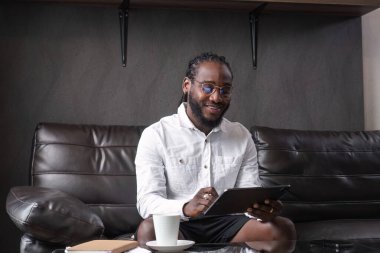 Genç Afrikalı Amerikalı bir adam evinden online olarak çalışıyor, modern bir oturma odasındaki kanepede otururken tablet kullanıyor..