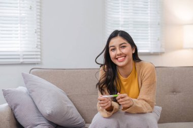 Hamilelik testi tutan neşeli bir yüzü olan genç bir kadın modern bir oturma odasında rahat bir kanepede oturuyor..