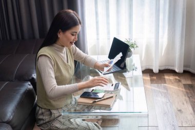 Evde çalışan genç bir kadın, dizüstü bilgisayar kullanıyor ve modern oturma odasındaki belgeleri inceliyor..