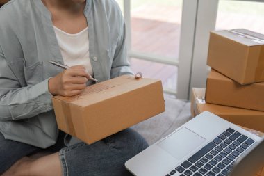 Küçük işletmesinde çalışan genç bir kadın dizüstü bilgisayar kullanırken kargo için paketler hazırlıyor..