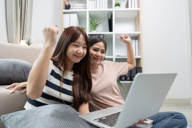 İki genç kadın online çalışmalardaki başarılarını kutluyorlar. Mutluluktan gülümseyip kollarını kaldırırken modern bir oturma odasında dizüstü bilgisayarlarıyla oturuyorlar..