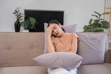 Başı ağrıyan bir kadın, modern bir oturma odasındaki bir koltukta oturuyor, başını rahatsızlık içinde tutuyor..