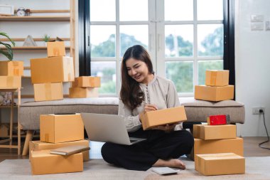Evde çalışan, internetten ürün satan genç bir kadın. Etrafı karton kutularla çevrili ve online mağazasını yönetmek için dizüstü bilgisayar kullanıyor..
