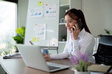 Bir kadın muhasebeci telefonda konuşarak ve bilgisayarını modern ofis ortamında çalıştırarak birden fazla iş yapar..