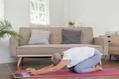 Evde yoga yapan yaşlı bir kadın, bilgisayarında online bir dersi takip ediyor, modern ve rahat bir oturma odasında..