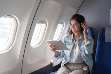 Uçaktaki genç iş kadını, bir tablet ve kulaklık kullanıyor, pencere kenarında oturuyor, modern hava yolculuğunu ve profesyonel yaşam tarzını temsil ediyor..