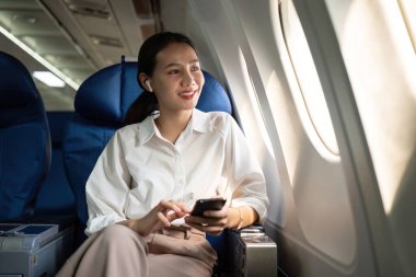 Genç bir muhasebeci uçakta çalışıyor, akıllı telefon kullanıyor ve kulaklık takıyor. İş seyahati ve modern teknoloji kavramı.