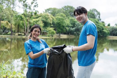 Parktaki gölün kenarındaki çöpleri temizlemeye gönüllü olan neşeli bir çift çevresel sorumluluk ve takım çalışması sergiliyor..
