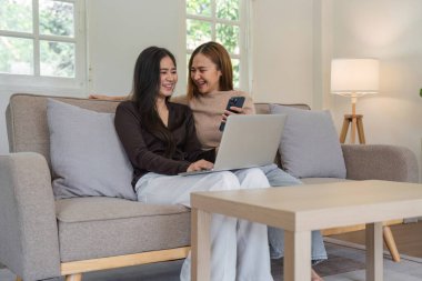 Kanepede oturan neşeli lezbiyen bir çift, dizüstü bilgisayar ve akıllı telefon kullanıyorlar, rahat, modern bir oturma odasında birlikte vakit geçiriyorlar..
