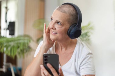 İletişim kurmak için akıllı telefon ve kulaklık kullanan, modern teknolojiyi ev ortamında sergileyen yaşlı bir kadın..