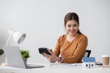 Bir kadın, modern bir ofiste masa başında oturarak, ev satın almak için finans planlarken hesap makinesi kullanır ve belgeleri gözden geçirir..