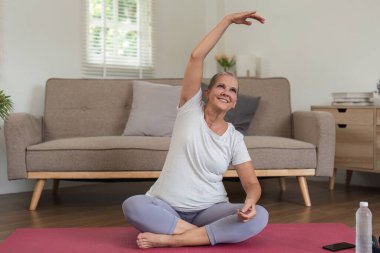 Yaşlı bir kadın evde yoga yapıyor, pembe bir paspasın üzerinde oturuyor, gülümsüyor ve modern mobilyalarla parlak bir oturma odasında esniyor..