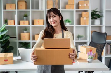 Ofisinde, online işi için kutu tutan genç bir kadının portresi. Arkaplanda paketler ve bitkiler olan raflar.