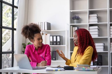 Genç Afrikalı Amerikalı iş kadını evrak işlerinden dolayı stresli hissediyor, modern ofis ortamında bir meslektaşıyla çözümleri tartışıyor..