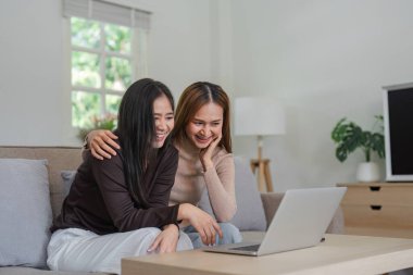 Evde dizüstü bilgisayar kullanan neşeli bir lezbiyen çift kanepede oturuyor. LGBT aşk, modern ilişkiler ve ev hayatı temaları için mükemmel..