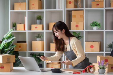 Modern bir ev ofisindeki genç bir kadın, nakliye kutuları ve ofis malzemeleriyle çevrili, e-ticaret ve küçük işletme operasyonları ile çevrimiçi ürün siparişlerini kontrol ediyor..