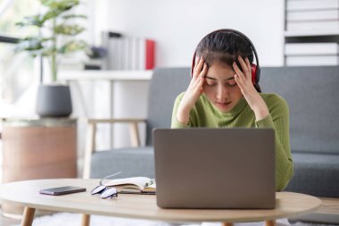 Genç bir kadın evde çalışırken stresli hissediyor. Dizüstü bilgisayarla, kulaklıkla ve modern oturma odasında bir defterle oturuyor..