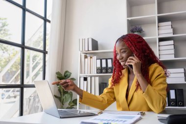 Kendine güvenen, sarı ceketli Afrikalı genç bir iş kadını modern bir ofiste oturuyor. Evrak işleri, dizüstü bilgisayar ve telefonla birden fazla iş yapıyor..