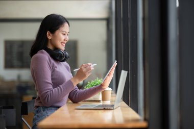 Modern bir kafede dizüstü bilgisayar ve tablet kullanan, kulaklık takan ve gülümseyen genç bir kadın..