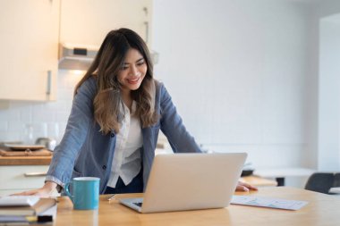 Modern bir mutfakta online olarak çalışan genç bir kadın, dizüstü bilgisayar kullanıyor ve bir fincan kahvenin tadını çıkarıyor, gülümsüyor ve işine odaklanıyor..