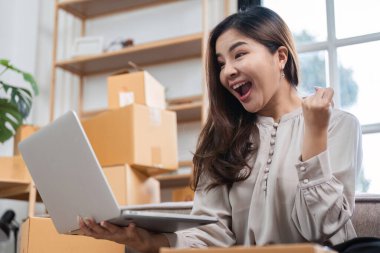 A young woman entrepreneur enthusiastically managing her online business, taking customer orders, and working on her laptop in a home office setting. clipart