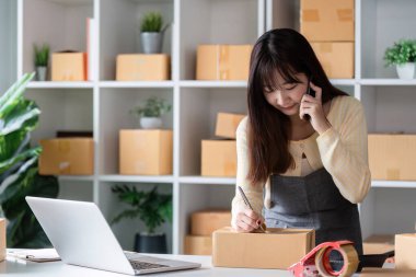 Modern bir ev ofisinde çalışan genç bir bayan girişimci, kutularla, dizüstü bilgisayarla ve telefonla online satışları yönetiyor..