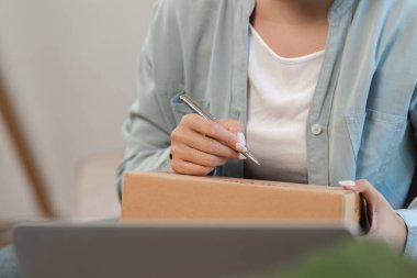 Genç bir kadın online satış işini yönetiyor, evde dizüstü bilgisayarla çalışırken bir paketin üzerine yazıyor..