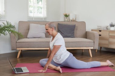 Elderly woman learning yoga online at home, following instructions on a laptop, promoting health and wellness in a modern living room setting. clipart