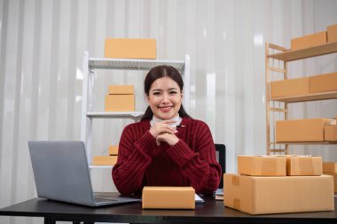 Evden çalışan gülümseyen bir kadın, internetten envanter kutuları ve modern bir ev ofisinde dizüstü bilgisayarla ürünler satıyor..