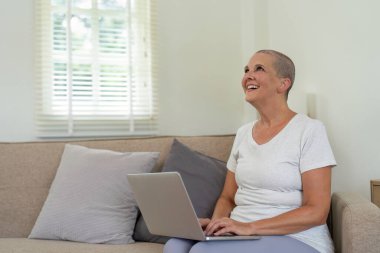 Kanepede dizüstü bilgisayar kullanarak oturan, gülümseyen ve modern teknolojiyle meşgul olan yaşlı bir kadın. Parlak ve rahat ev içi doğal ışıkla dolu..