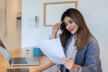 Evden bağımsız olarak çalışan genç bir kadın dizüstü bilgisayar kullanıyor ve modern ofis ortamında belgeleri inceliyor..