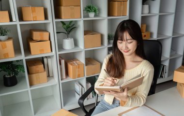 Modern bir ev ofisinde çalışan genç bir bayan girişimci, çevresindeki kutular ve envanterlerle online satışları yönetiyor..