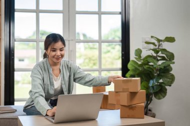 Evinden internet üzerinden satış yapan, dizüstü bilgisayar kullanan ve teslimat için paketleri düzenleyen genç bir kadın..