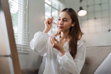 Beyaz bluzlu bir kadın sabahları cilt bakımı yapıyor. Parlak ve modern bir iç mekanda oturuyor..