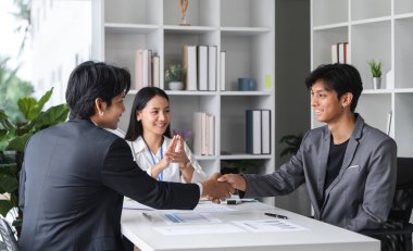 İki işadamı el sıkışma ve sözleşme imzalama ile bir anlaşma imzalarken, bir iş kadını modern bir ofiste onaylayıcı bir şekilde bakıyor.