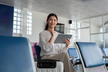 Havaalanı terminalinde oturan bir kadın, elinde bir tablet, uçağa binmeyi bekliyor. Modern seyahat ve teknoloji kavramı.