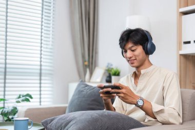 Genç bir adam modern bir oturma odasında, kulaklık takıyor ve akıllı telefon kullanıyor, boş zamanlarının keyfini çıkarıyor..