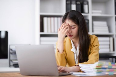Modern bir ofiste genç bir iş kadını kendini stresli ve çalışmaktan bunalmış hissediyor, bir masa başında dizüstü bilgisayar ve bir yığın evrakla oturuyor..