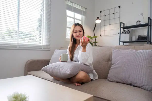 stock image A woman relaxes on a sofa with a cup of coffee and an apple, enjoying her morning routine in a bright, modern living room.