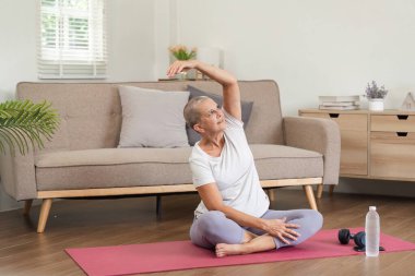 Yaşlı bir kadın, modern oturma odasındaki bir paspasın üzerinde rahatlatıcı bir yoga pozu veriyor. Sağlık ve sağlığını destekliyor..