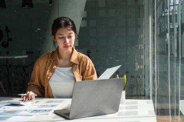 Genç bir iş kadını, parlak ve modern bir ofis ortamında dizüstü bilgisayarı üzerinde çalışırken belgeleri analiz etmeye odaklanır..