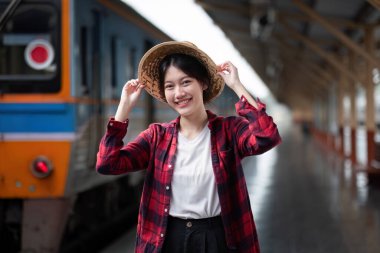 Sevimli genç bir kadın tren istasyonunda duruyor, gülümsüyor ve treni bekliyor. Şapka ve günlük kıyafetler giyiyor, neşeli ve modern bir atmosfer yaratıyor..